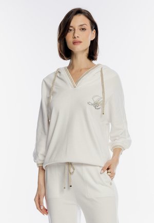 Kobieta ubrana w biały długi rękaw z kapturem z złotą lamówką i dopasowane białe spodnie, stojąca z jedną ręką w kieszeni na jednolitym tle.