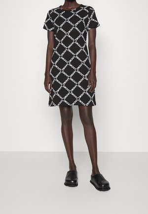 Robe noire à manches courtes avec motif diagonal blanc du logo DKNY, portée avec des chaussures noires slip-on, debout devant un fond uni.