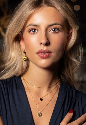 Gold jewelry displayed includes three layered necklaces: thin chains with circular pendants and a textured, sun-like earring. Dark top.