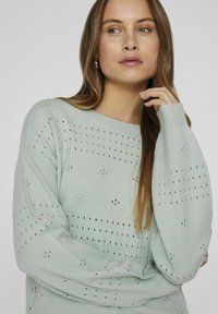 Světl zelený pletený svetr s volným střihem, zdobený dekorativními dírkovými vzory a žebrovanými manžetami, kulatým výstřihem a měkkou texturou.