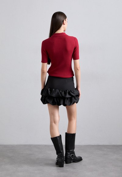 Femme aux longs cheveux noirs se tient face à un mur gris, portant un top rouge à manches courtes côtelé, une mini jupe noire à volants et des bottes noires mi-mollet.