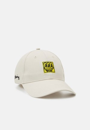 Beżowa czapka baseballowa z zakrzywionym daszkiem, z żółtą naszywką z uśmiechniętą, trzyoczyną twarzą z przodu i czarnym napisem z boku.