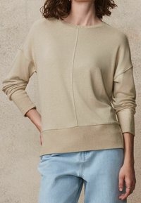 Pull beige à manches longues avec un col rond, des poignets et un ourlet côtelés. Présente un détail de couture subtil sur le devant. Associé à un jean bleu clair.