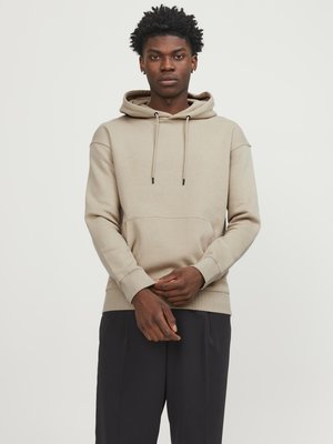 Haut à capuche beige avec cordon de serrage et poche avant, présentant des manches longues et des poignets côtelés. Porté avec un pantalon sombre ample.