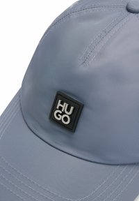 Gorra azul claro hecha de tela suave, con un parche cuadrado negro que lleva "HUGO" en texto plateado y un detalle de costura en el visor.