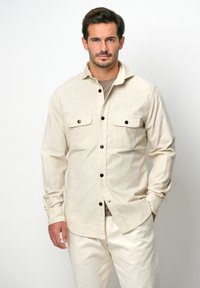 Lichtbeige button-up shirt met twee borstzakken, zwarte knopen en een zachte textuur, gecombineerd met lichtcrème broeken.
