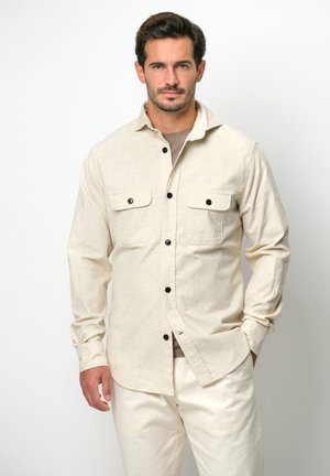 Lyse beige skjorte med knapper og to brystlommer, sorte knapper og en blød tekstur, parret med lys cremefarvede bukser.