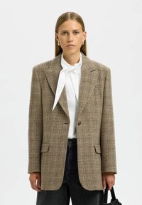 Blazer à carreaux brun avec une coupe structurée, revers crantés, fermeture à un bouton et poches à l'avant. Porté sur un blouse blanche et un jean noir.