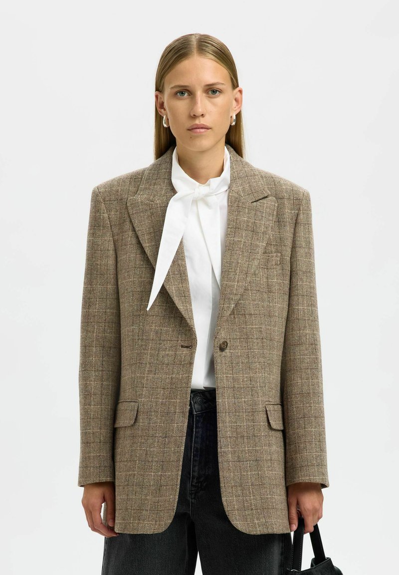 Blazer à carreaux brun avec une coupe structurée, revers crantés, fermeture à un bouton et poches à l'avant. Porté sur un blouse blanche et un jean noir.