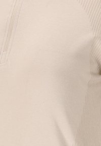 Lys beige top med et tekstureret ribbet design langs skuldrene og en halv-zip halsudskæring; blødt, glat stof med en tætsiddende pasform.