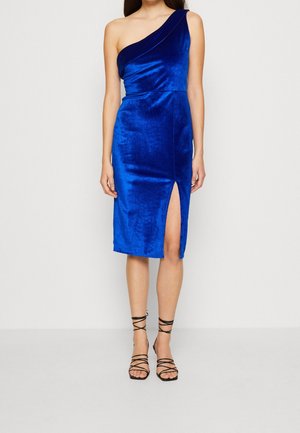 Mujer con vestido de terciopelo azul real hasta la rodilla, con un solo hombro, abertura alta en el muslo y sandalias negras de tacón alto con tiras.