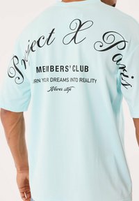 Homme portant un t-shirt bleu clair avec un texte noir au dos disant "Club des membres Project X Paris Transformez vos rêves en réalité Ne jamais s'arrêter."
