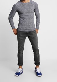 Langärmeliges marineblaues Strickshirt mit Textur, kombiniert mit grauen Jeans aus Denim und blauen Sneakers mit weißen Akzenten. Lässige Passform; hochgekrempelte Jeanssäume.