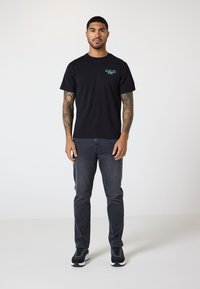 Svart bomull t-skjorte med frontgrafisk logo, kombinert med mørkegrå jeans og svarte sneakers. Modellen står mot en ensfarget bakgrunn.