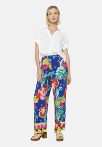 Bela srajca s gumbi v kombinaciji z živopisnimi modrimi hlačami s cvetličnim vzorcem, ki vključujejo zelene liste, rožnate in rumene poudarke ter sproščen kroj.