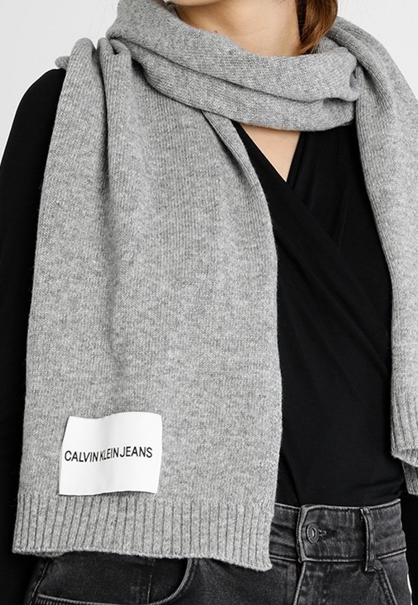 Szary dziergany szalik z metką Calvin Klein Jeans noszony na czarnej bluzce i ciemnych dżinsach, pokazujący górną część tułowia i szyję.