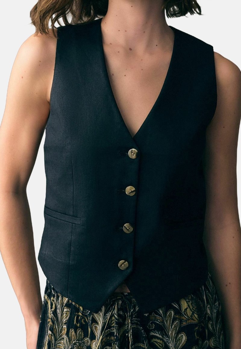 Femme portant un gilet noir ajusté sans manches boutonné sur un pantalon à motifs feuillus, visible des épaules jusqu’aux hanches.