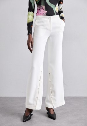 Femme portant un pantalon blanc à jambes larges avec des détails de boutons le long des jambes avant, associé à un haut noir à fleurs à manches longues et des chaussures noires pointues.