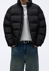 Personne portant une veste matelassée noire par-dessus un sweat-shirt gris clair, un jean bleu, une ceinture noire avec des œillets argentés, et un collier chaîne en argent.