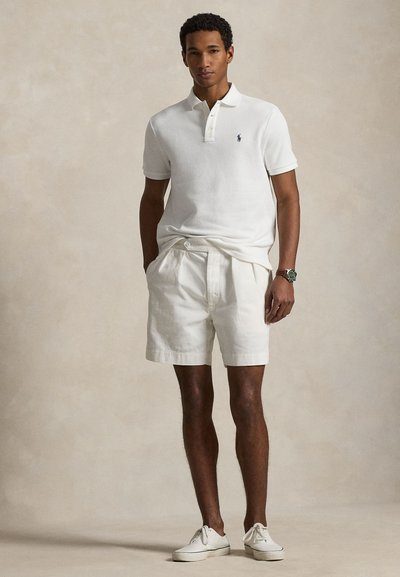Mężczyzna w białej koszulce polo z krótkim rękawem, białych szortach, białych trampkach i zegarku na nadgarstku, stojący z rękami w kieszeniach.