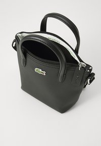Sac à main noir texturé avec doubles poignées supérieures et fermeture éclair. Présente un logo crocodile vert et une bandoulière détachable pour plus de polyvalence.
