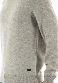 Zachte lichtgrijze gebreide trui met geribbelde onderkant en manchetten, voorzien van een klein donker rechthoekig etiket van stof nabij de onderkant.