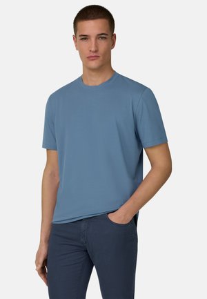 Jeune homme portant un t-shirt bleu uni à manches courtes et un pantalon bleu foncé, debout avec une main dans la poche devant un fond uni.