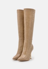 Bottes beige montant au genou avec un motif texturé effet crocodile, bout pointu et talon aiguille, présentant une finition lisse et un design élégant.
