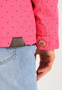 Giacca rosa con pois neri, con un'etichetta in pelle marrone e dettagli con bottoni di legno. Indossata sopra denim azzurro chiaro.