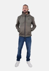 Giacca con cappuccio in tessuto verde oliva con due tasche sul petto, chiusura con zip e una texture liscia, abbinata a jeans blu e sneakers bianche.