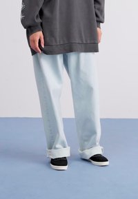 Lichtblauwe, opgerolde jeans gecombineerd met een oversized sweatshirt in antracietgrijs en zwarte sneakers met witte zolen op een blauwe achtergrond.