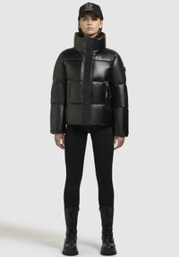 Zwarte pufferjack met hoge kraag, glanzende textuur en gewatteerd ontwerp. Gecombineerd met zwarte leggings en hoge laarzen met veters.