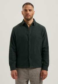 Man in een donkergroen textuur shirt met knopen en een patroon van bruine en beige broek, staand tegen een effen lichte achtergrond.