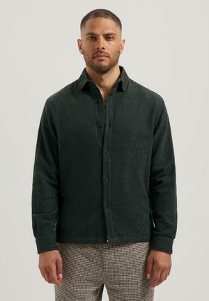 Man wearing a dark green textured button-up shirt and patterned brown and beige pants, standing against a plain light background.