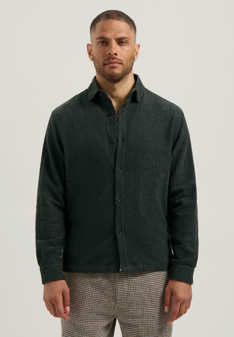 Man in een donkergroen textuur shirt met knopen en een patroon van bruine en beige broek, staand tegen een effen lichte achtergrond.
