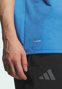 T-shirt in cotone blu con colletto rotondo, dotato di branding "climaCOOL" vicino all'orlo. Pantaloni neri con logo Adidas grigio sulla coscia.