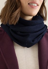 Écharpe en tricot bleu marine avec une texture subtile et des perles bleues éparpillées, portée sur un pull en tricot beige à torsades et un manteau bordeaux.