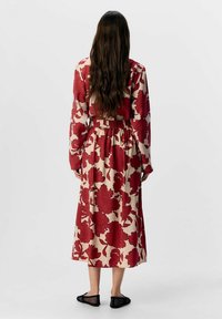 Langærmet kjole med et rødt blomstermønster på en beigefarvet baggrund, med samlet talje og længde til midt på læggen. Kombineret med sorte slip-on sko.