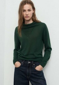 Pull en maille verte avec un col montant, des poignets côtelés et une texture douce, associé à un jean en denim bleu foncé.