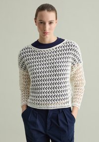 Maglione bianco all'uncinetto con motivo a maglia aperta, vestibilità rilassata, scollatura ampia, indossato sopra una maglietta girocollo blu navy, abbinato a pantaloni scuri.
