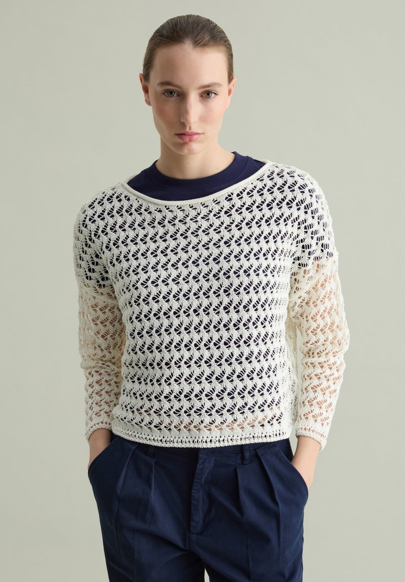 Maglione bianco all'uncinetto con motivo a maglia aperta, vestibilità rilassata, scollatura ampia, indossato sopra una maglietta girocollo blu navy, abbinato a pantaloni scuri.
