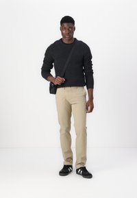Pull en maille noire, pantalon beige coupe droite, baskets noires avec des rayures blanches, et un sac à épaule noir. Design simple et fonctionnel.