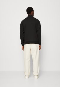 Sweatshirt noir avec un col montant, des poignets et un ourlet côtelés. Porté avec un pantalon crème à coupe décontractée et des baskets blanches.