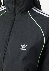 Svart vindjacka med hög krage, med vita tre-stripes-accenter och grönt kantband. Inkluderar en broderad logotyp på bröstet.