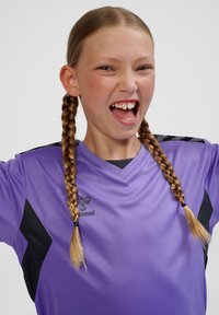 Lila sportströja med svarta detaljer, korta ärmar och V-ringning. Modellen har flätat hår och ler med ett entusiastiskt uttryck.