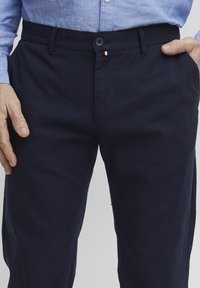 Pantaloni in cotone blu navy con patta con bottone, tasche frontali e un piccolo accento tricolore vicino alla vita. Texture morbida.