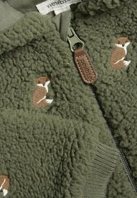 Olijfgroene fleecjas met rits, ribgebreide mouwen en kleine bruine geborduurde dierenpatronen. Zachte textuur met een knusse uitstraling.