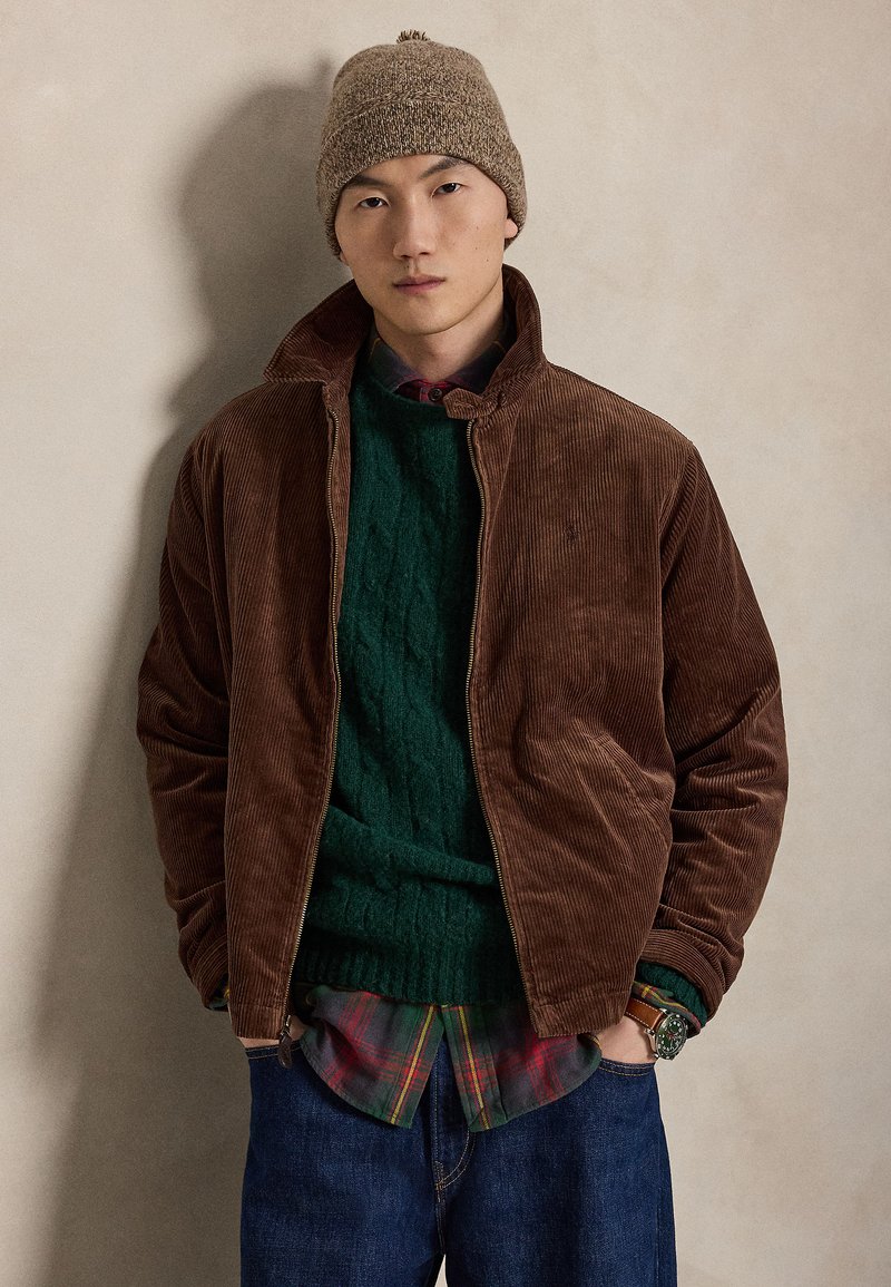 Ung mand iført en brun kordfløjlsjakke, grøn sweater, ternet skjorte, blå jeans, brun strikhue og armbåndsur står foran en neutral baggrund.