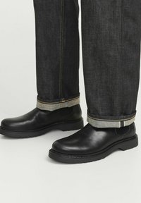 Botas de cuero negras hasta el tobillo con puntera redonda y suela de goma, combinadas con unos vaqueros de mezclilla oscuros que tienen un dobladillo enrollado y costuras en contraste.