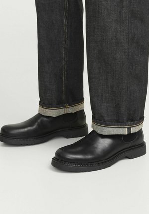 Botas de cuero negras hasta el tobillo con puntera redonda y suela de goma, combinadas con unos vaqueros de mezclilla oscuros que tienen un dobladillo enrollado y costuras en contraste.